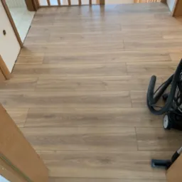 Ułożone panele podłogowe w korytarzu, widoczne przejście schodowe i odkurzacz, jasne drewno.