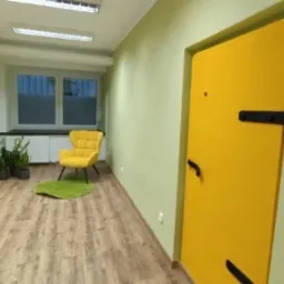 Jasne biuro z drewnianą podłogą, zielonymi ścianami i żółtymi akcentami: fotel, drzwi i dywanik w kształcie liścia.