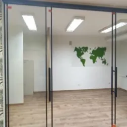 Przeszklone drzwi przesuwne z czarnymi ramami i uchwytami, oddzielające biuro z tapetą w palmy od drugiego pomieszczenia z mapą świata na ścianie i drewnianą podłogą.