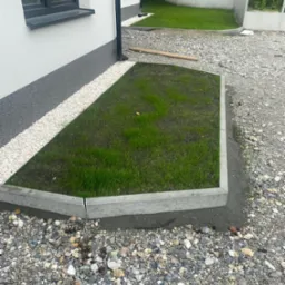 Narożny, wykończony klomb z młodą trawą przy białym domu. Obrzeże z betonowych elementów. Szara nawierzchnia z kamyków. Widok na fragment trawnika i betonowy mur.