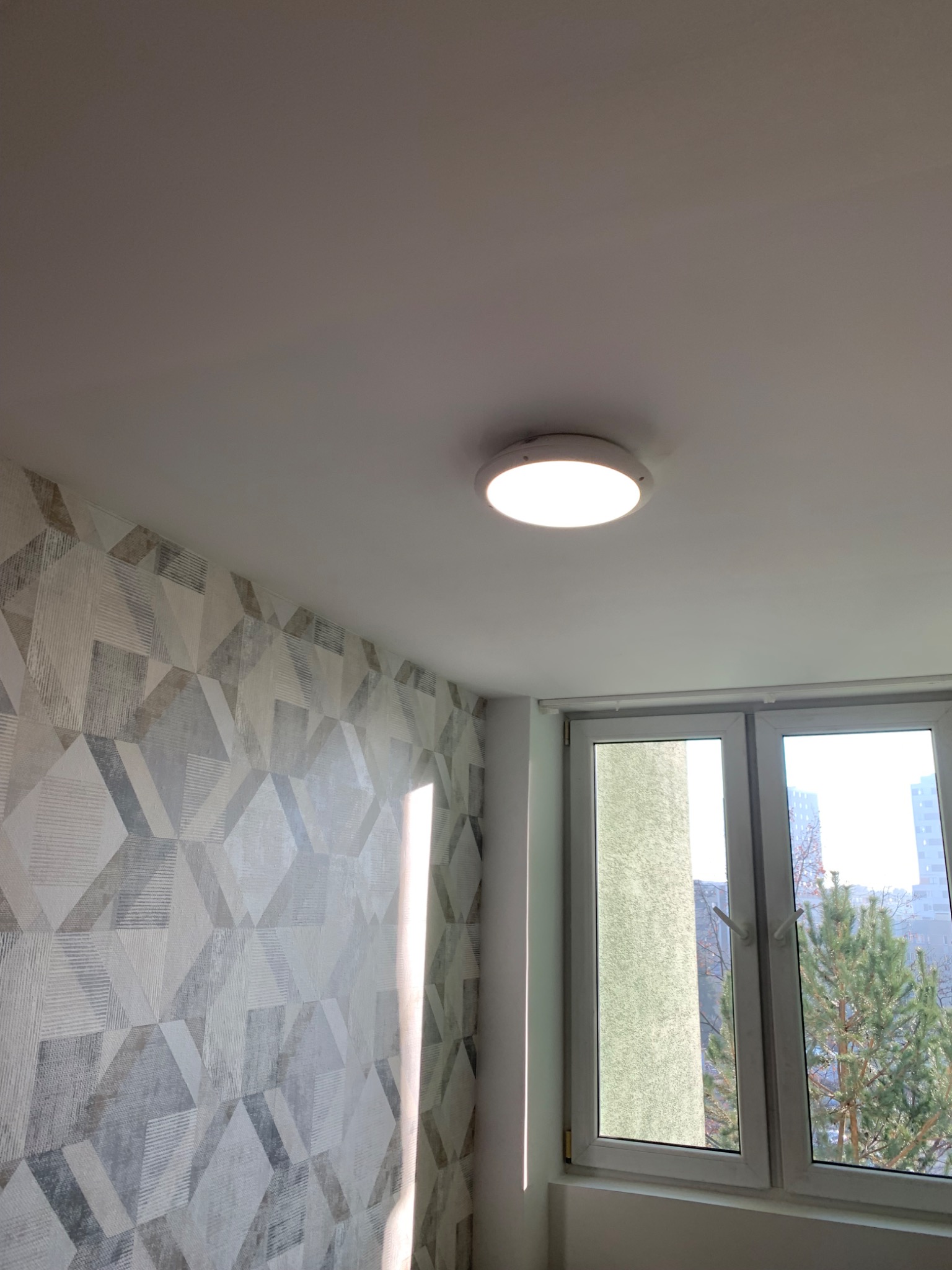 Wnętrze pokoju po remoncie: ściana z geometryczną tapetą w odcieniach szarości, biały sufit z okrągłą lampą LED i okno z widokiem na drzewo i budynki.