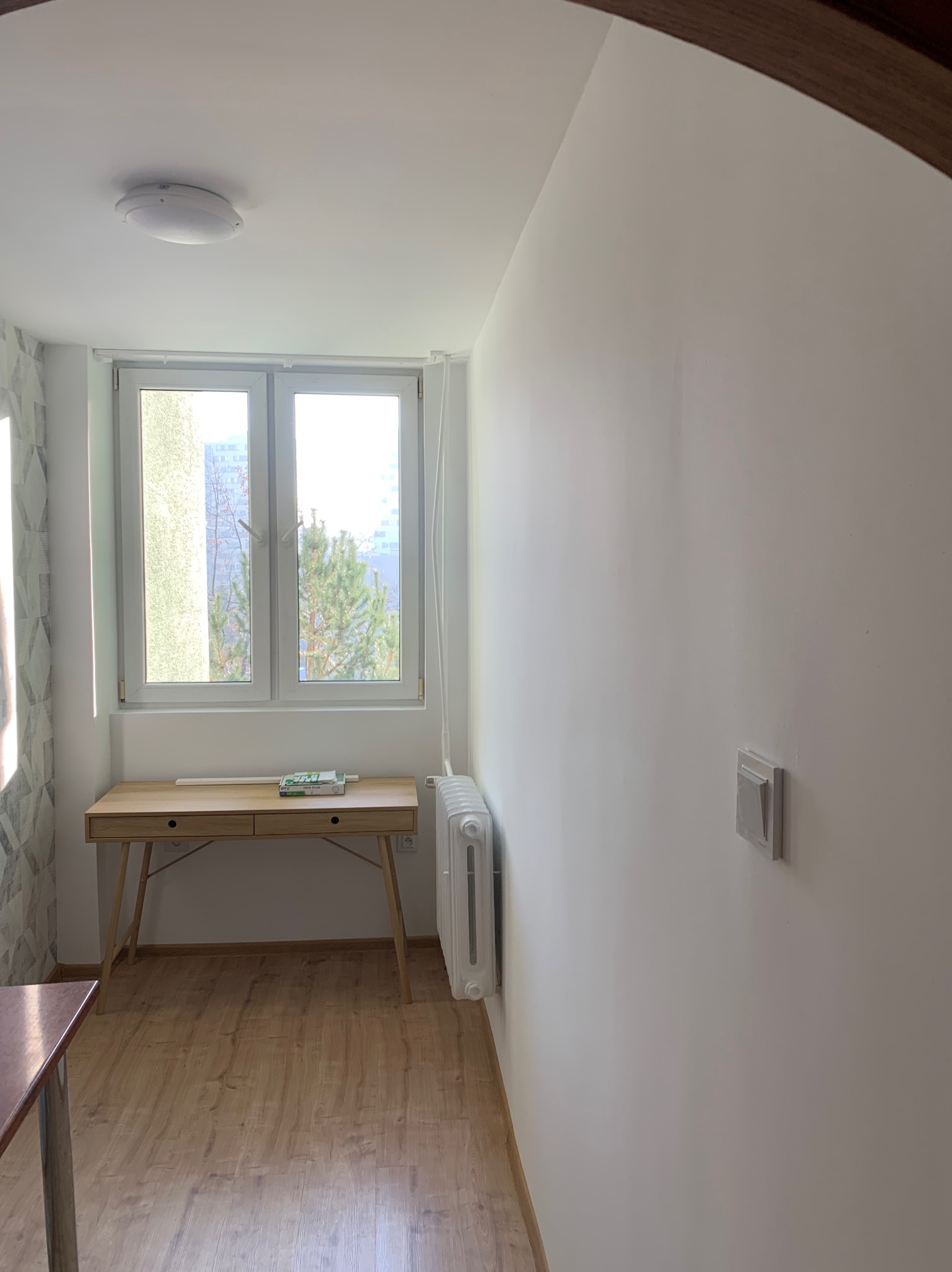 Pomalowane na biało wnętrze pokoju z drewnianą podłogą i biurkiem pod oknem, widoczny fragment ściany z włącznikiem, grzejnik i fragment innego mebla.