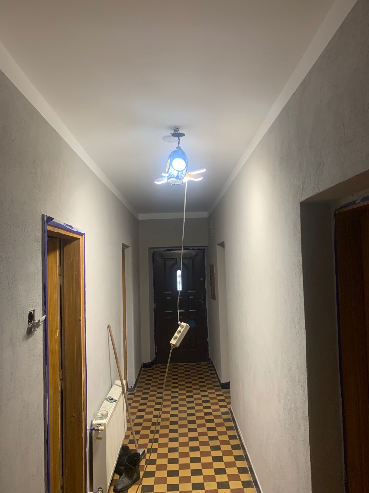 Korytarz w trakcie remontu z szarą, chropowatą fakturą ścian, podłogą w szachownicę, prowizorycznym oświetleniem w postaci lampy podłączonej do przedłużacza wiszącego z sufitu i zabezpieczonymi...