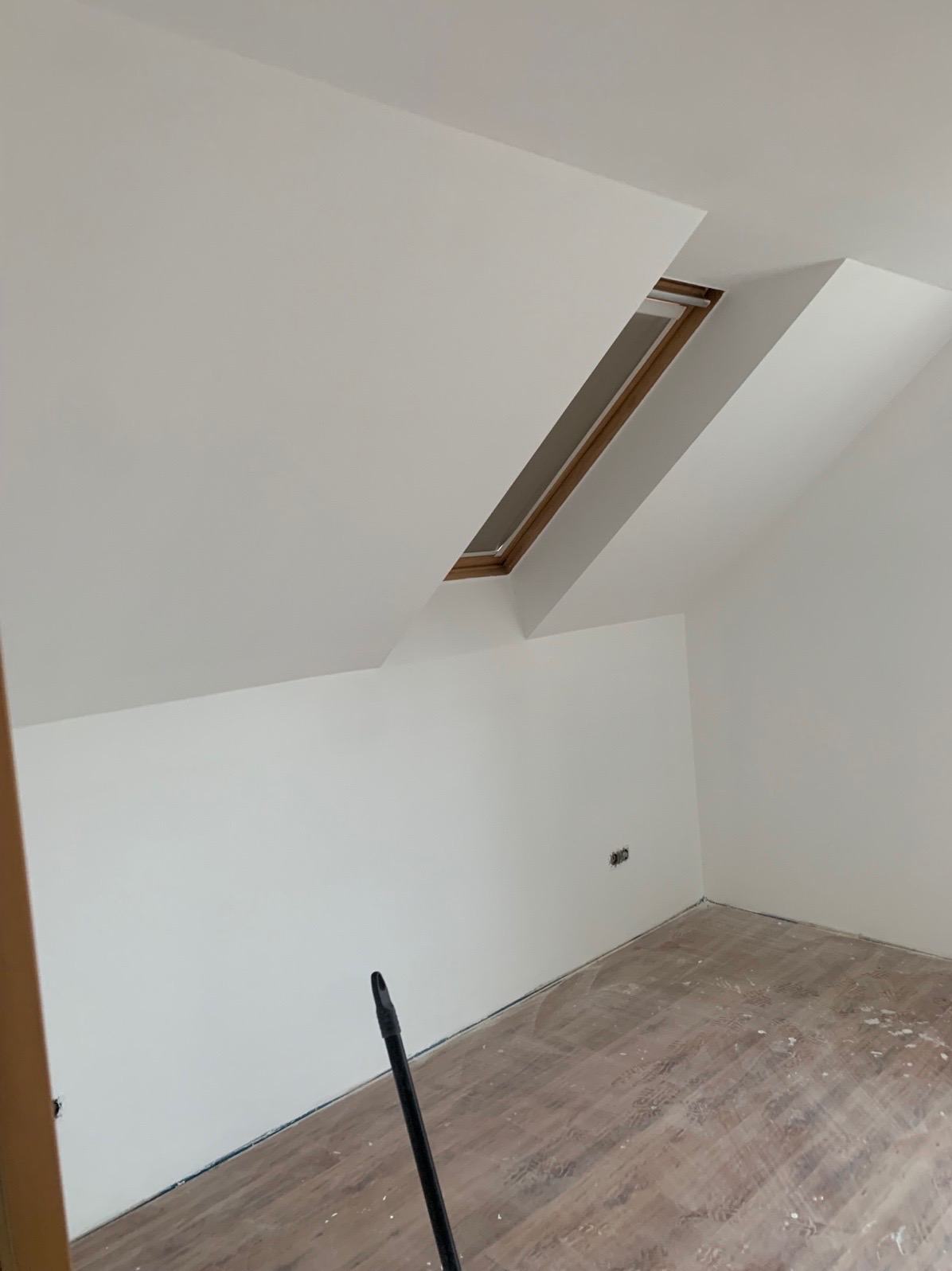 Poddasze po malowaniu na biało, widoczne okno dachowe, ślady farby na panelach podłogowych i wystające gniazdko elektryczne.