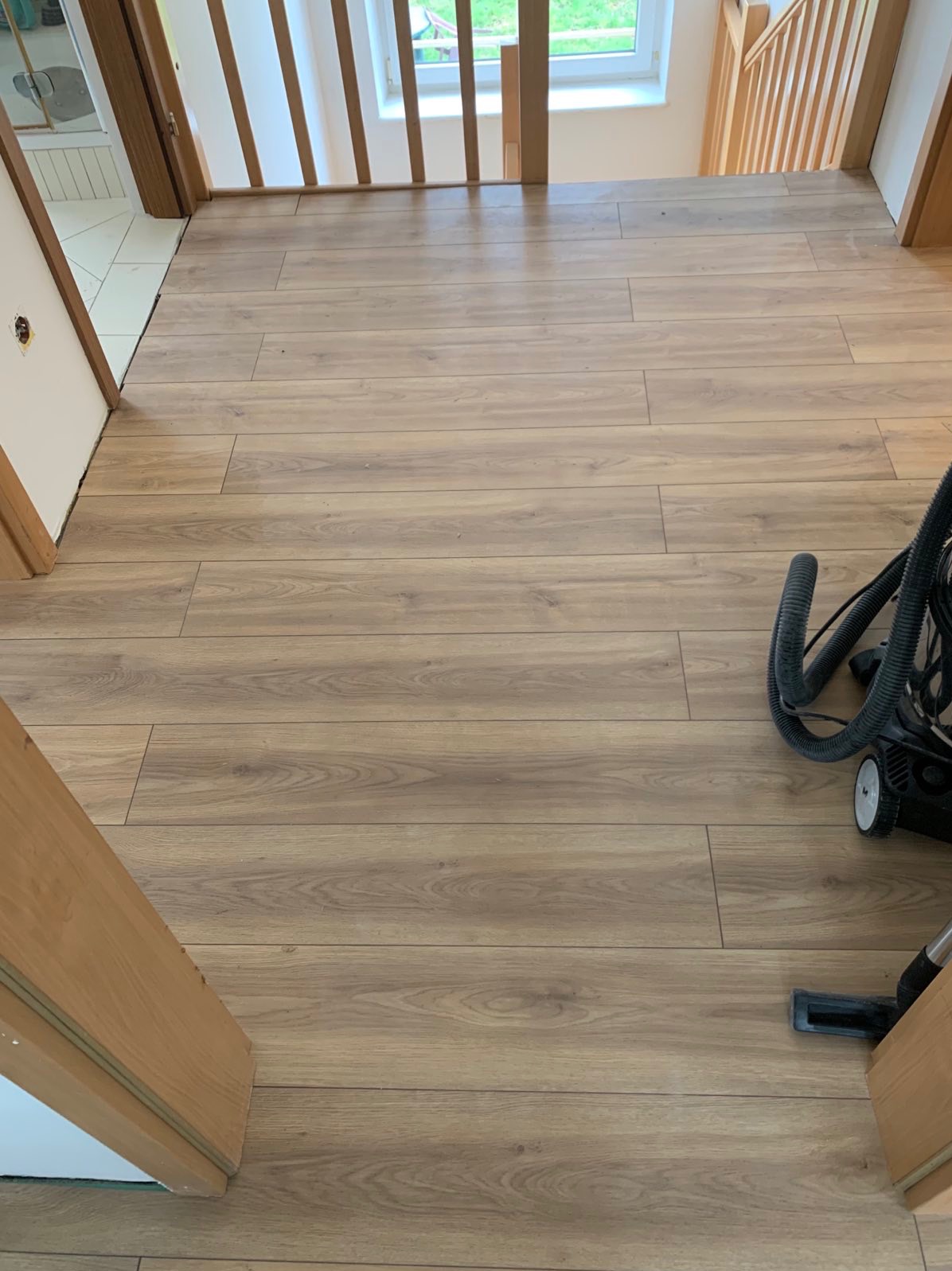 Ułożone panele podłogowe w korytarzu, widoczne przejście schodowe i odkurzacz, jasne drewno.