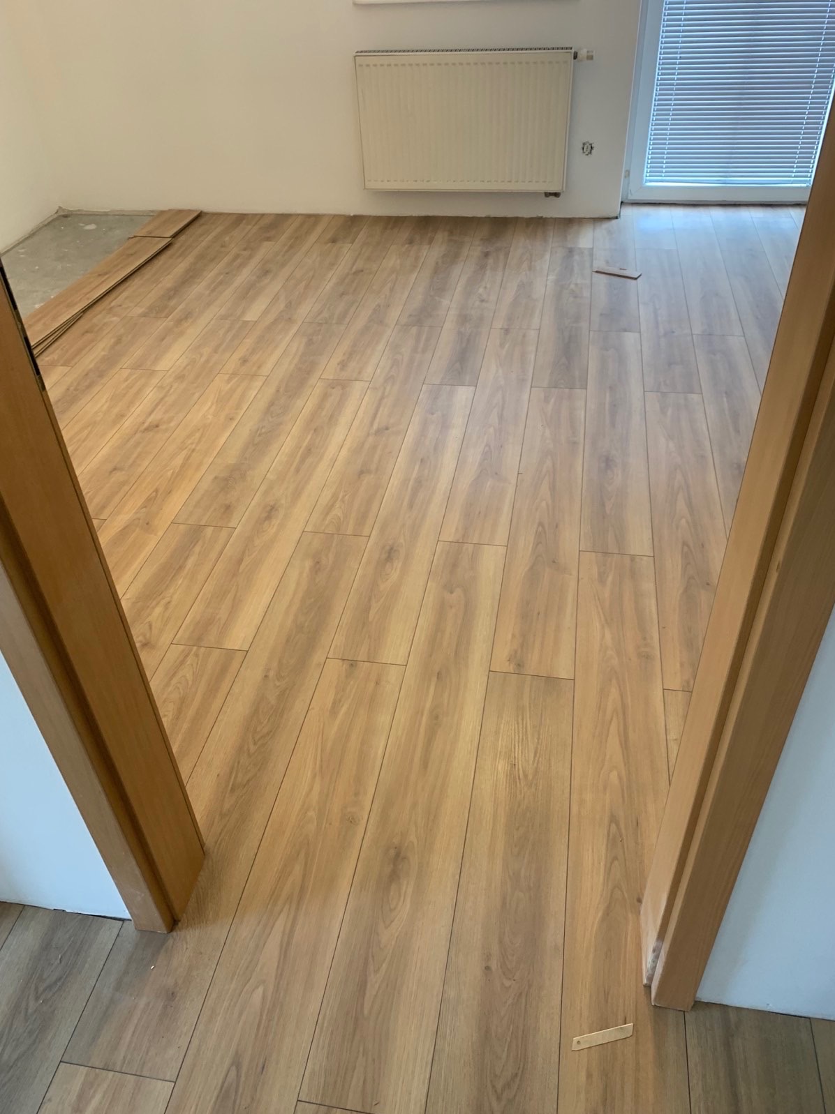Jasne panele podłogowe z widoczną strukturą drewna, ułożone w nowym mieszkaniu, widok z perspektywy wejścia przez drewniane futryny, fragment nieułożonych paneli w rogu pomieszczenia.