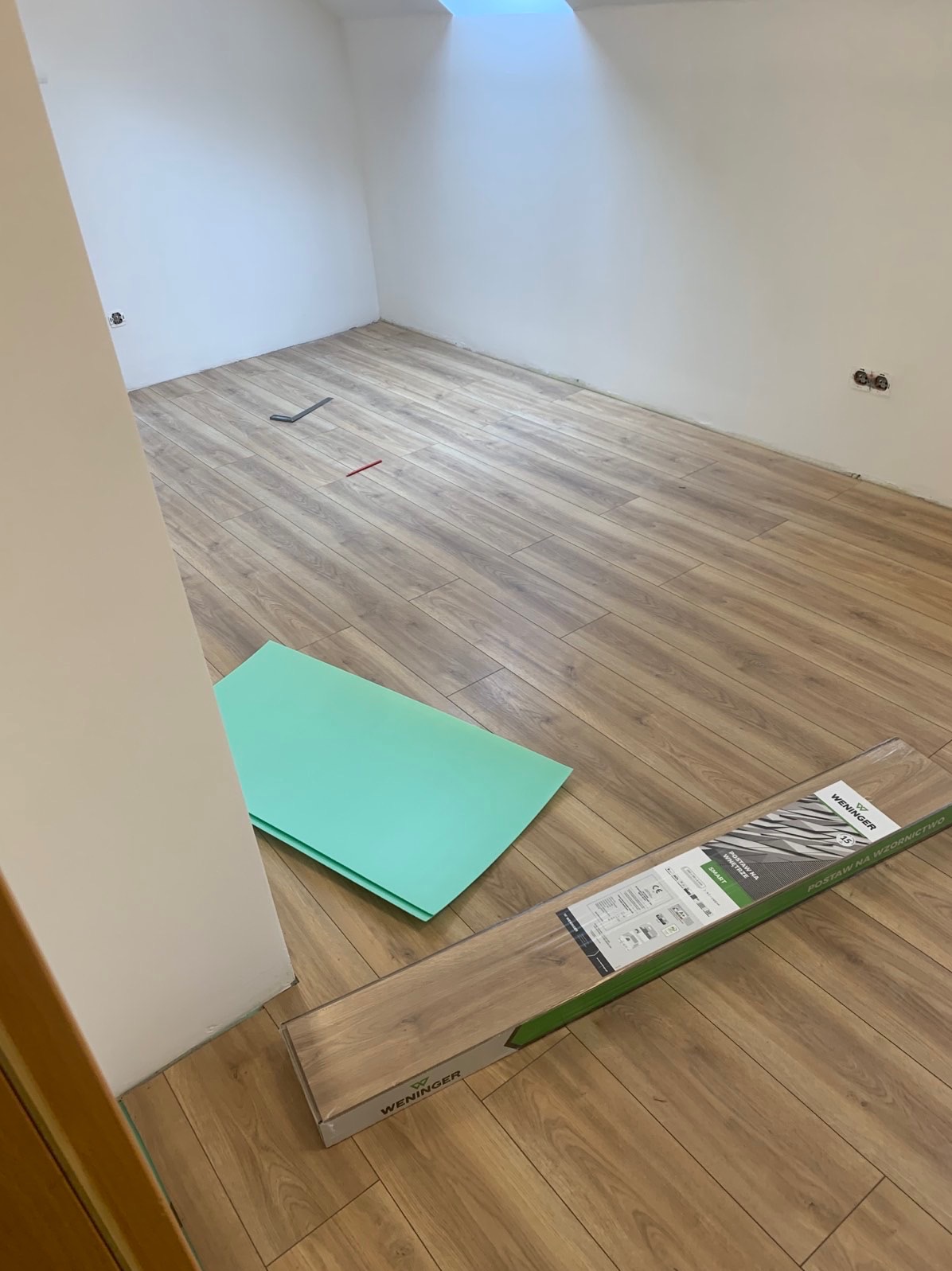 Pokój na poddaszu z częściowo ułożonymi panelami podłogowymi, widoczna paczka paneli marki Wenninger, zielona pianka pod panele, ołówek i narzędzie do montażu na podłodze.