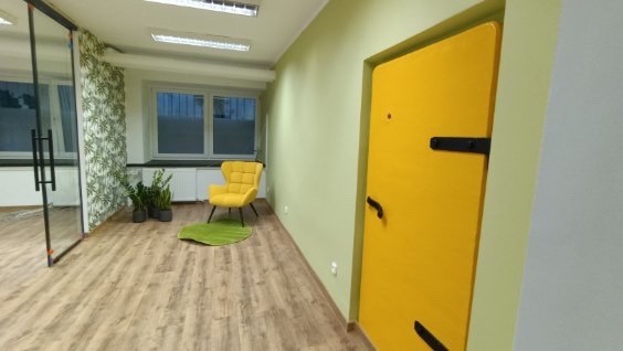 Jasne biuro z drewnianą podłogą, zielonymi ścianami i żółtymi akcentami: fotel, drzwi i dywanik w kształcie liścia.