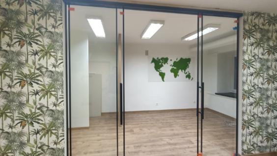 Przeszklone drzwi przesuwne z czarnymi ramami i uchwytami, oddzielające biuro z tapetą w palmy od drugiego pomieszczenia z mapą świata na ścianie i drewnianą podłogą.