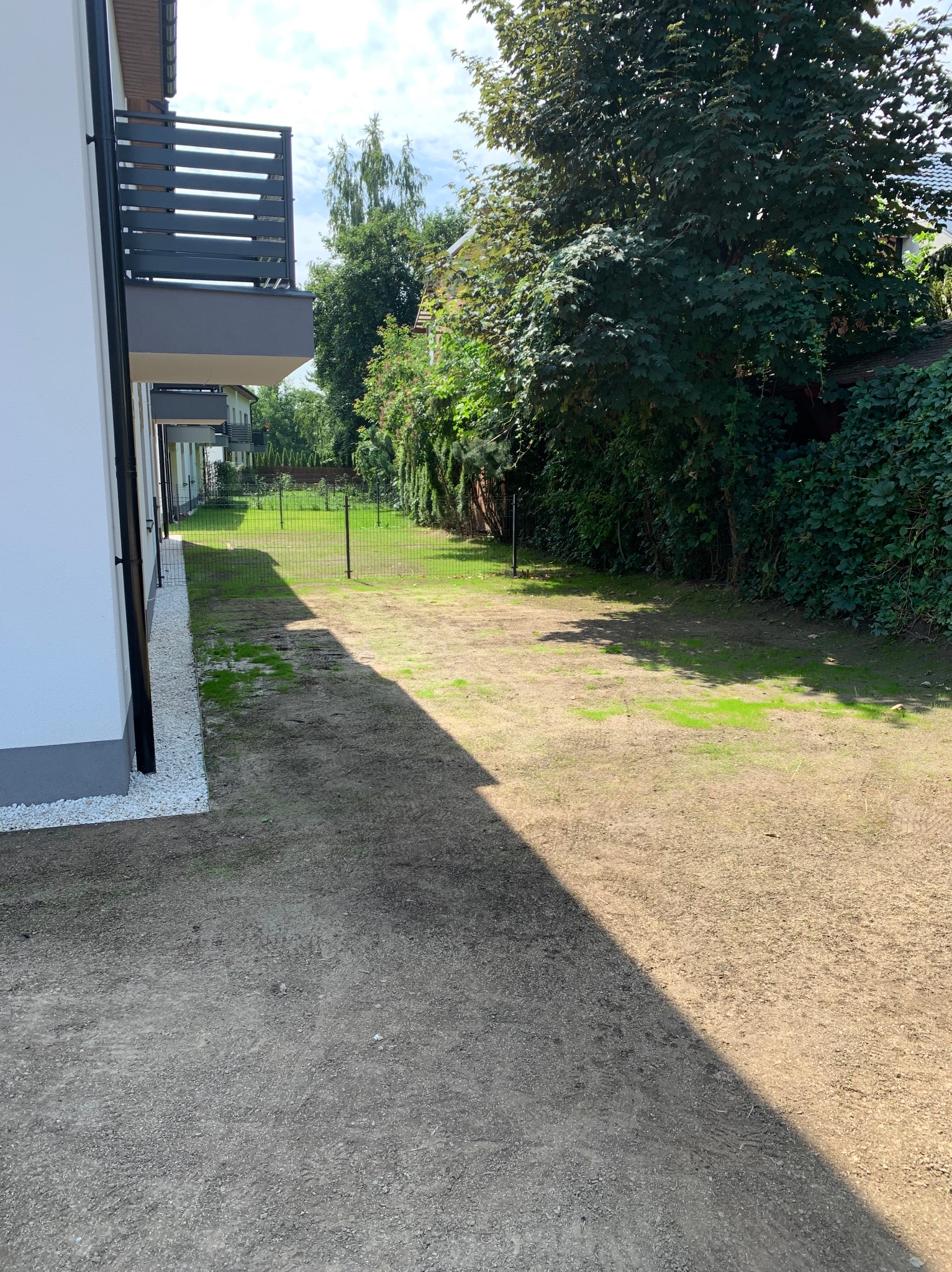 Widok z boku budynku na przygotowany teren pod trawnik, z widocznymi fragmentami trawy i ogrodzeniem w tle. Białe kamienie dekoracyjne przy elewacji.