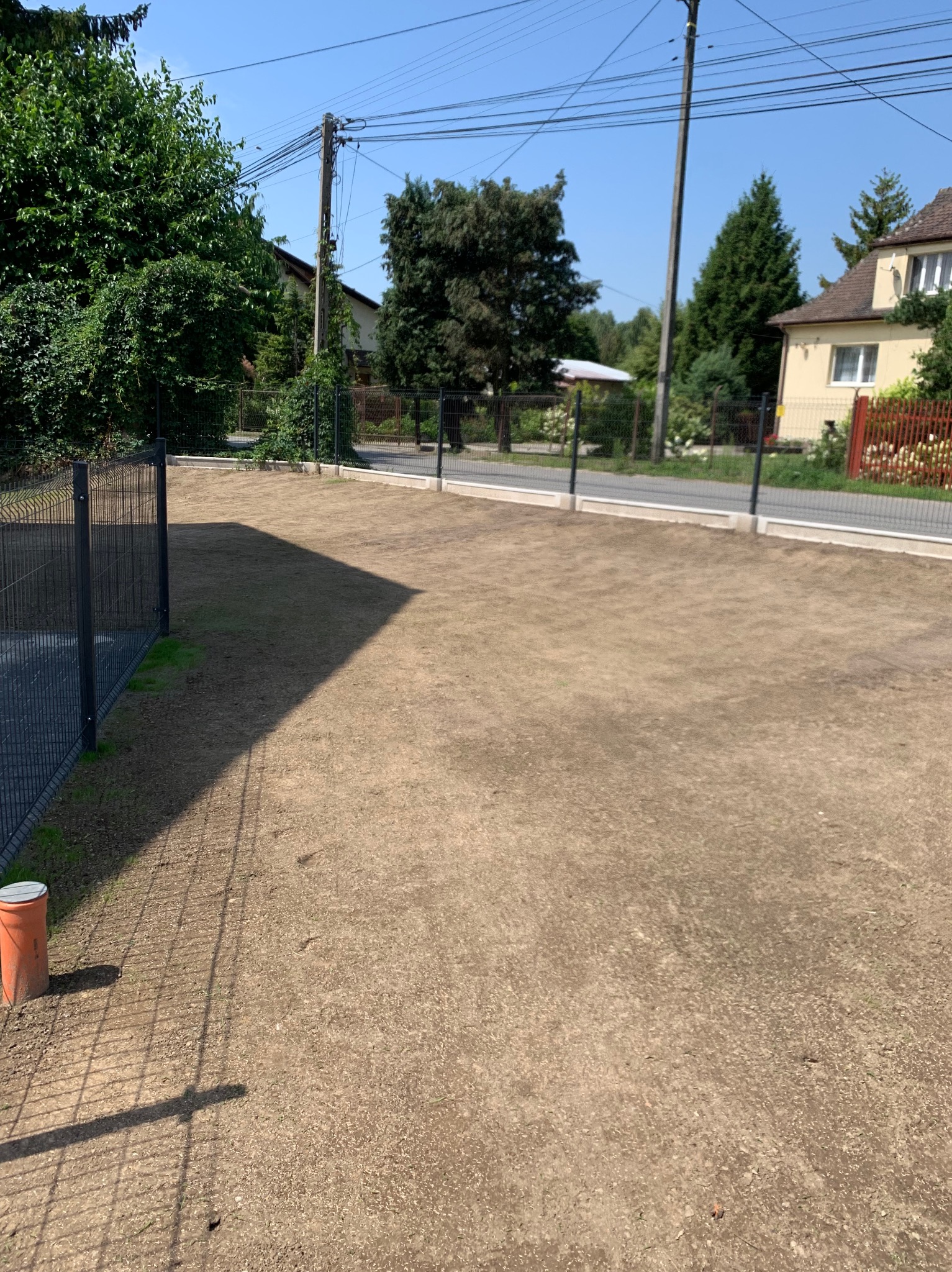 Przygotowany teren pod trawnik, widoczny system nawadniania. Grunt wyrównany, ogrodzony siatką. W tle domy i zieleń.