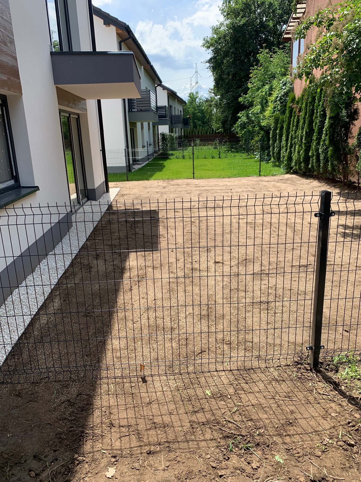 Działka z ogrodzeniem panelowym, przygotowana pod trawnik. Widoczny budynek z białymi ścianami i szarymi elementami architektonicznymi. Słoneczny dzień.