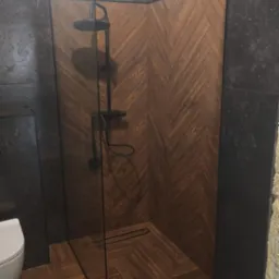 Nowoczesna kabina prysznicowa z drewnopodobnymi płytkami ułożonymi w jodełkę na ścianach i podłodze, czarna armatura, szklana ściana kabiny z czarną ramą, fragment toalety widoczny po lewej stronie.