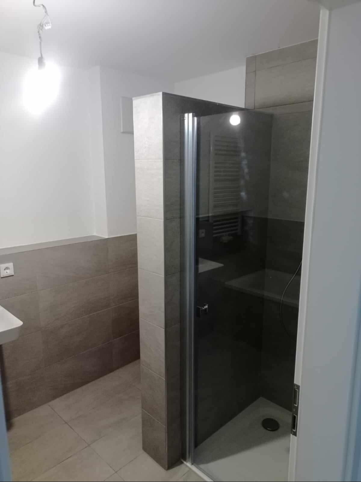 Nowoczesna kabina prysznicowa z szarymi płytkami i chromowanymi detalami, widoczny grzejnik drabinkowy wewnątrz kabiny, surowa żarówka na suficie.