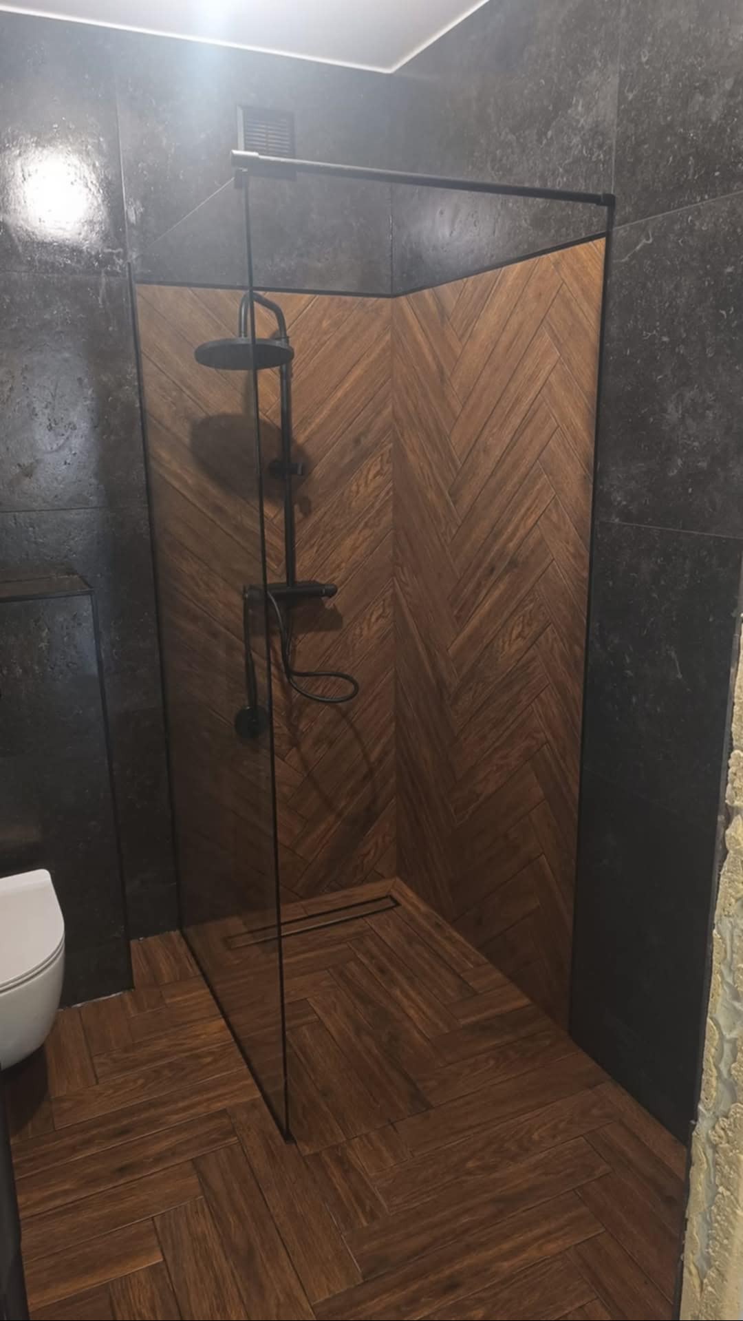 Nowoczesna kabina prysznicowa z drewnopodobnymi płytkami ułożonymi w jodełkę na ścianach i podłodze, czarna armatura, szklana ściana kabiny z czarną ramą, fragment toalety widoczny po lewej stronie.