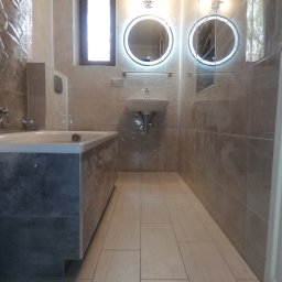 Tantum Optima - Nowoczesna łazienka z beżowymi płytkami, podświetlanymi okrągłymi lustrami, wanną obudowaną płytkami w odcieniach szarości i jasną podłogą z płytek imitujących drewno.