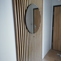 Tantum Optima - Drewniana ścianka działowa z pionowych listew z okrągłym lustrem, odbijającym drzwi, na tle jasnej ściany i podłogi.