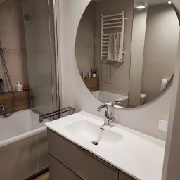 Tantum Optima - Nowoczesna łazienka z okrągłym lustrem, białą umywalką na szafce w kolorze taupe, kabiną prysznicową ze szklanymi drzwiami i grzejnikiem drabinkowym z ręcznikiem.