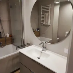 Nowoczesna łazienka z okrągłym lustrem, białą umywalką na szafce w kolorze taupe, kabiną prysznicową ze szklanymi drzwiami i grzejnikiem drabinkowym z ręcznikiem.