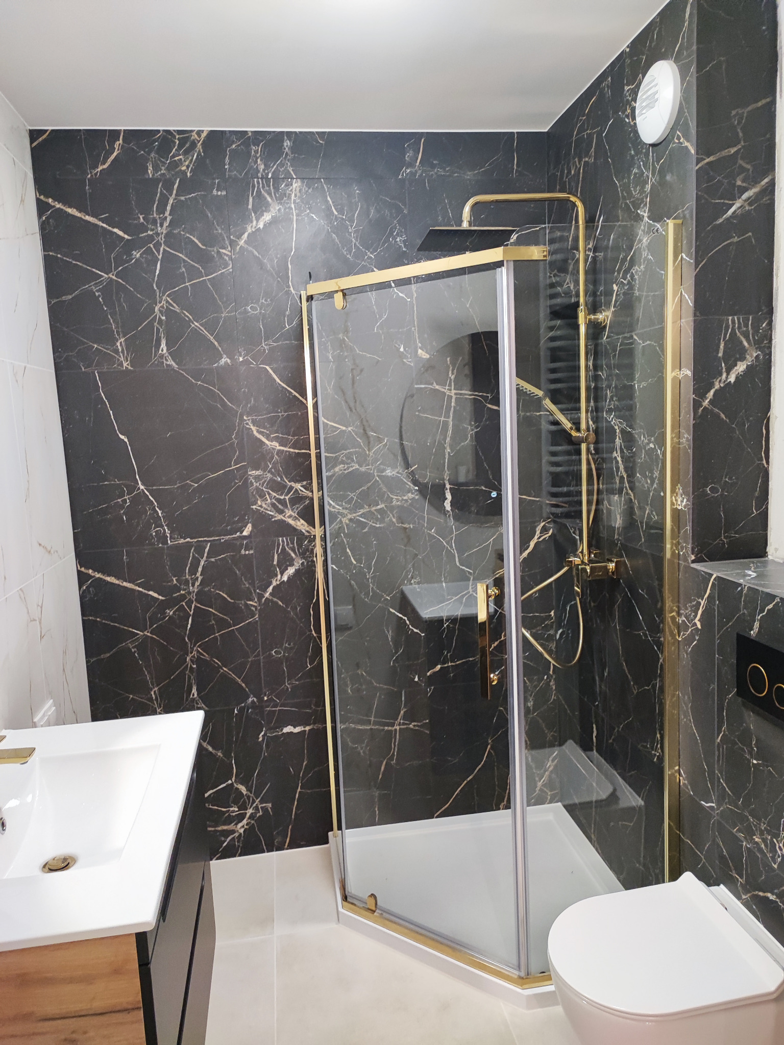 Nowoczesna łazienka z kabiną prysznicową w złotych ramach, czarnymi płytkami imitującymi marmur z białymi żyłami, białą umywalką na drewnianej szafce i białą toaletą.