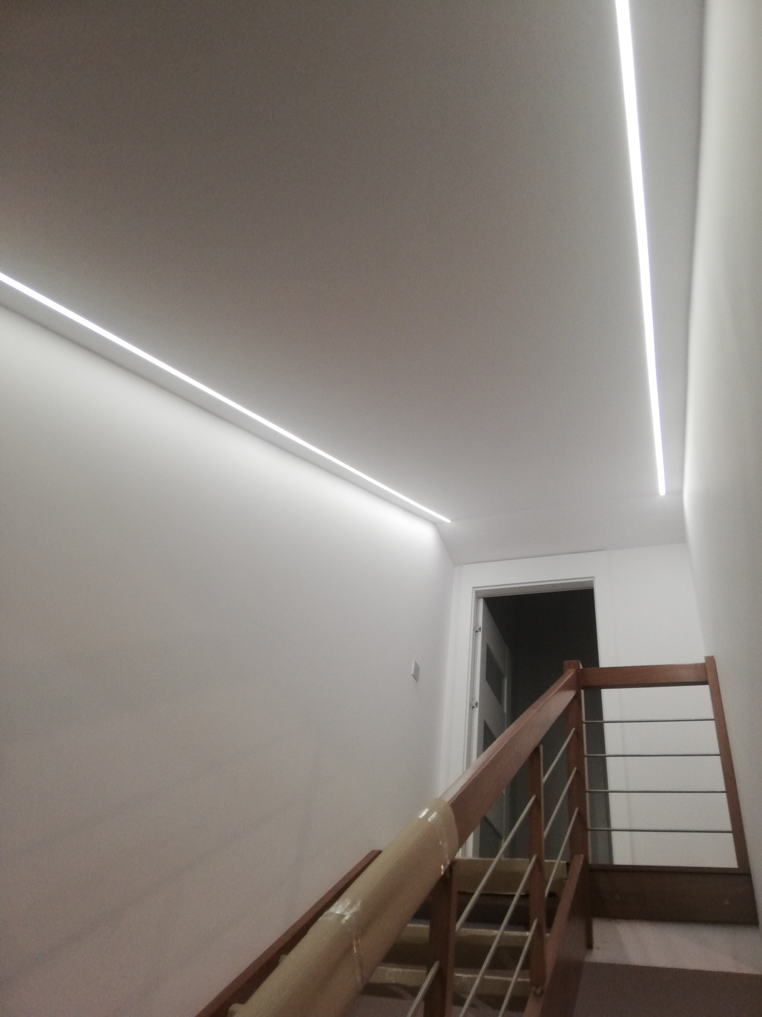 Korytarz ze schodami oświetlony dwoma liniami LED w suficie, drewniane balustrady schodów z metalowymi prętami, drzwi w tle.