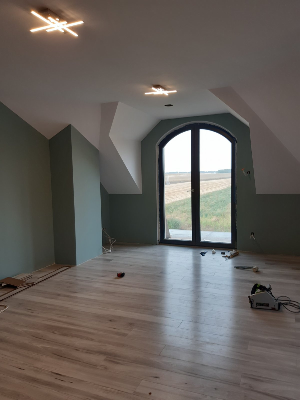 Pokój na poddaszu z drewnopodobną podłogą, ściany w odcieniach zieleni, duże okno łukowe z widokiem na pole, nowoczesne lampy sufitowe i rozłożone narzędzia do układania podłogi.