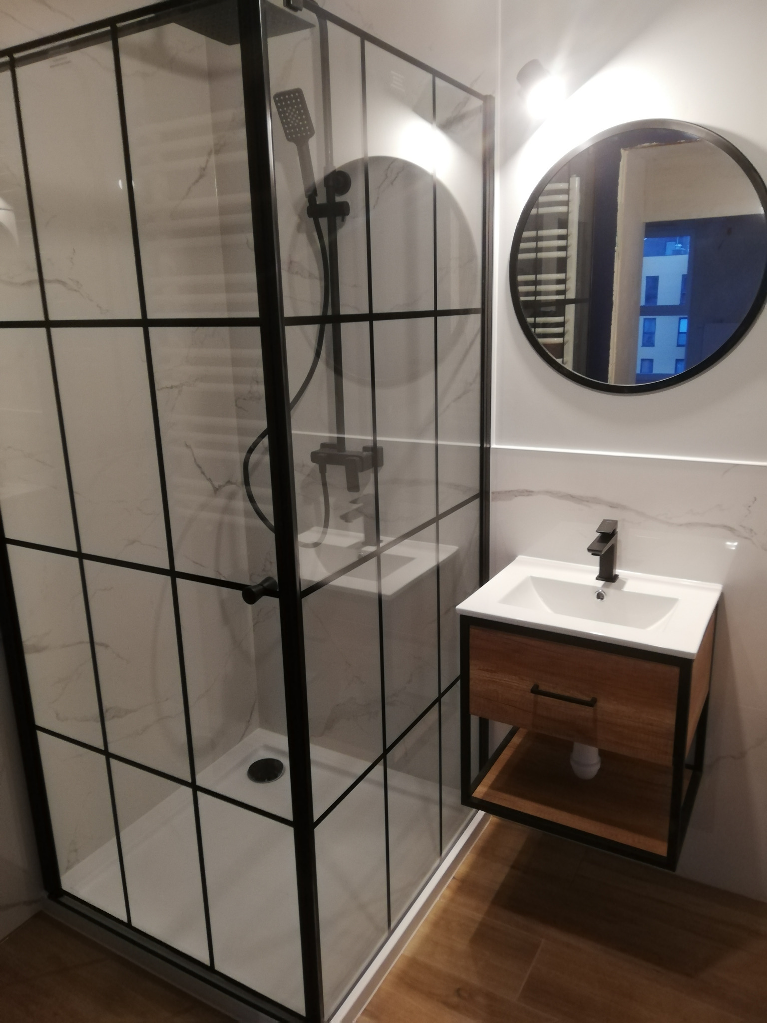 Nowoczesna łazienka z kabiną prysznicową typu walk-in z czarnymi szprosami i szafką pod umywalkę z drewnianym frontem oraz okrągłym lustrem.