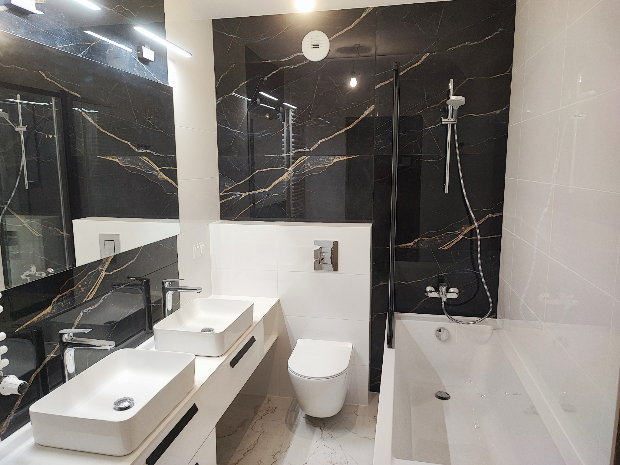Nowoczesna łazienka z czarnymi płytkami imitującymi marmur z złotymi żyłami, białą armaturą, dwiema umywalkami nablatowymi, wiszącą toaletą i kabiną prysznicową ze szklaną ścianką.