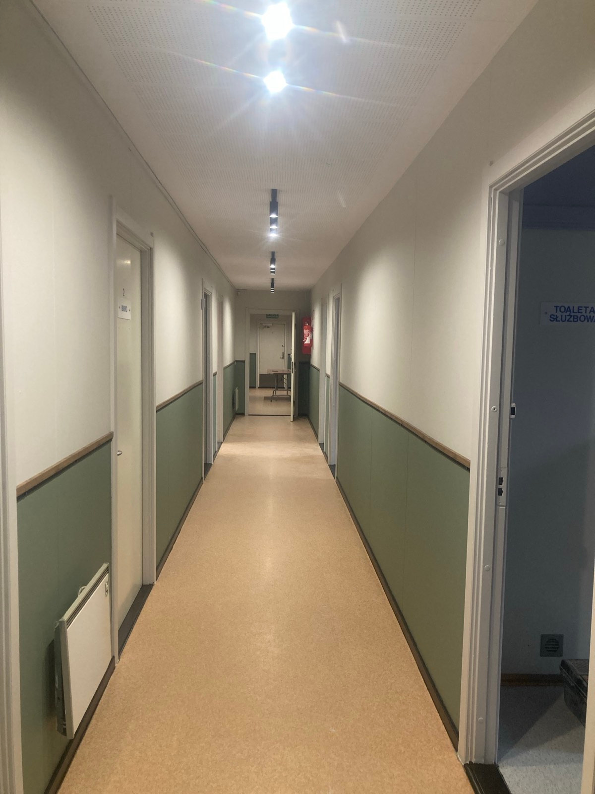 Długi korytarz biurowy z panelami ściennymi w kolorze oliwkowym, oświetleniem punktowym i drzwiami po obu stronach, widok w perspektywie.