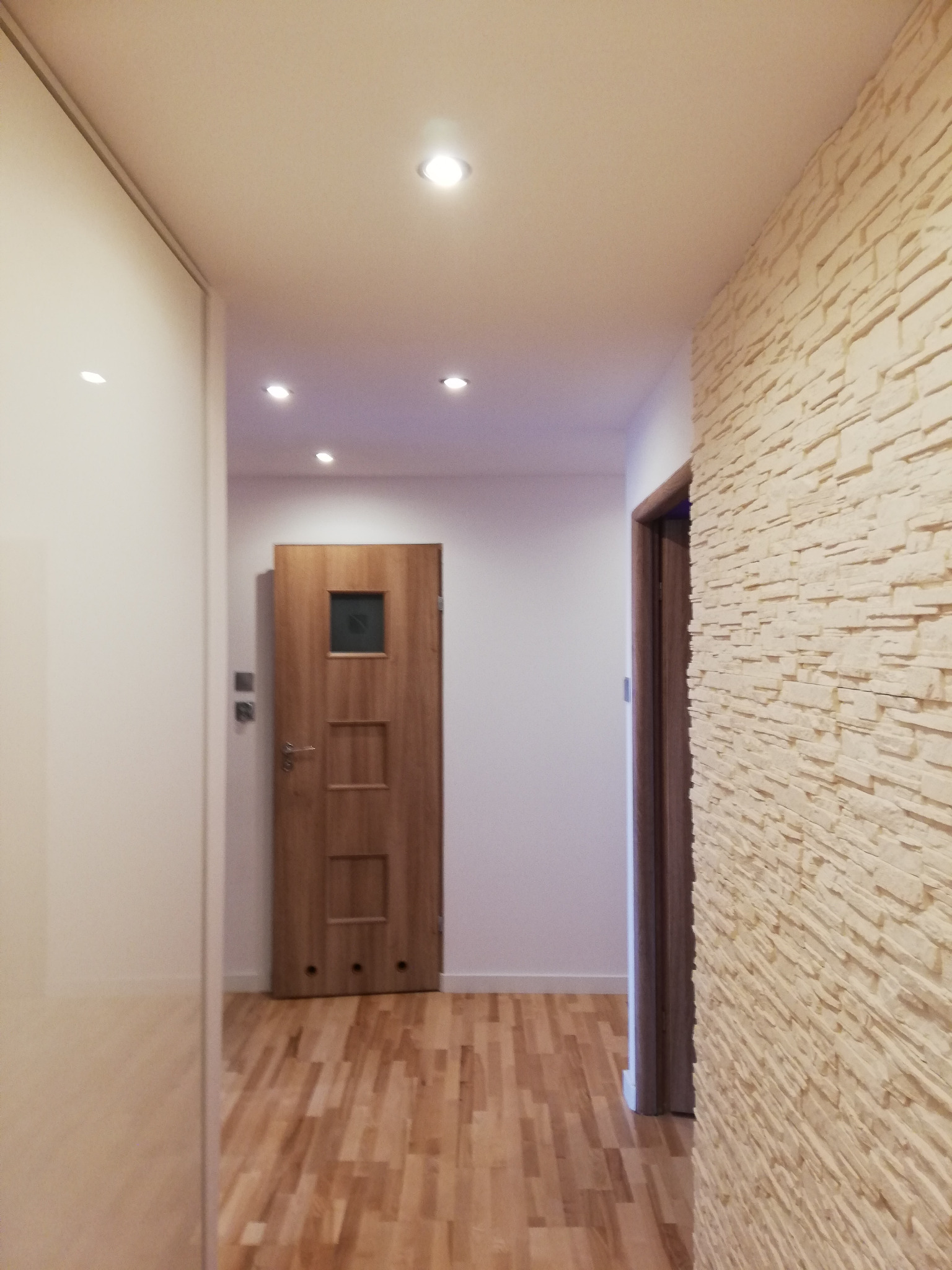 Korytarz po remoncie z drewnianą podłogą, ścianą wyłożoną dekoracyjnym kamieniem i dwiema drewnianymi drzwiami z przeszkleniami.