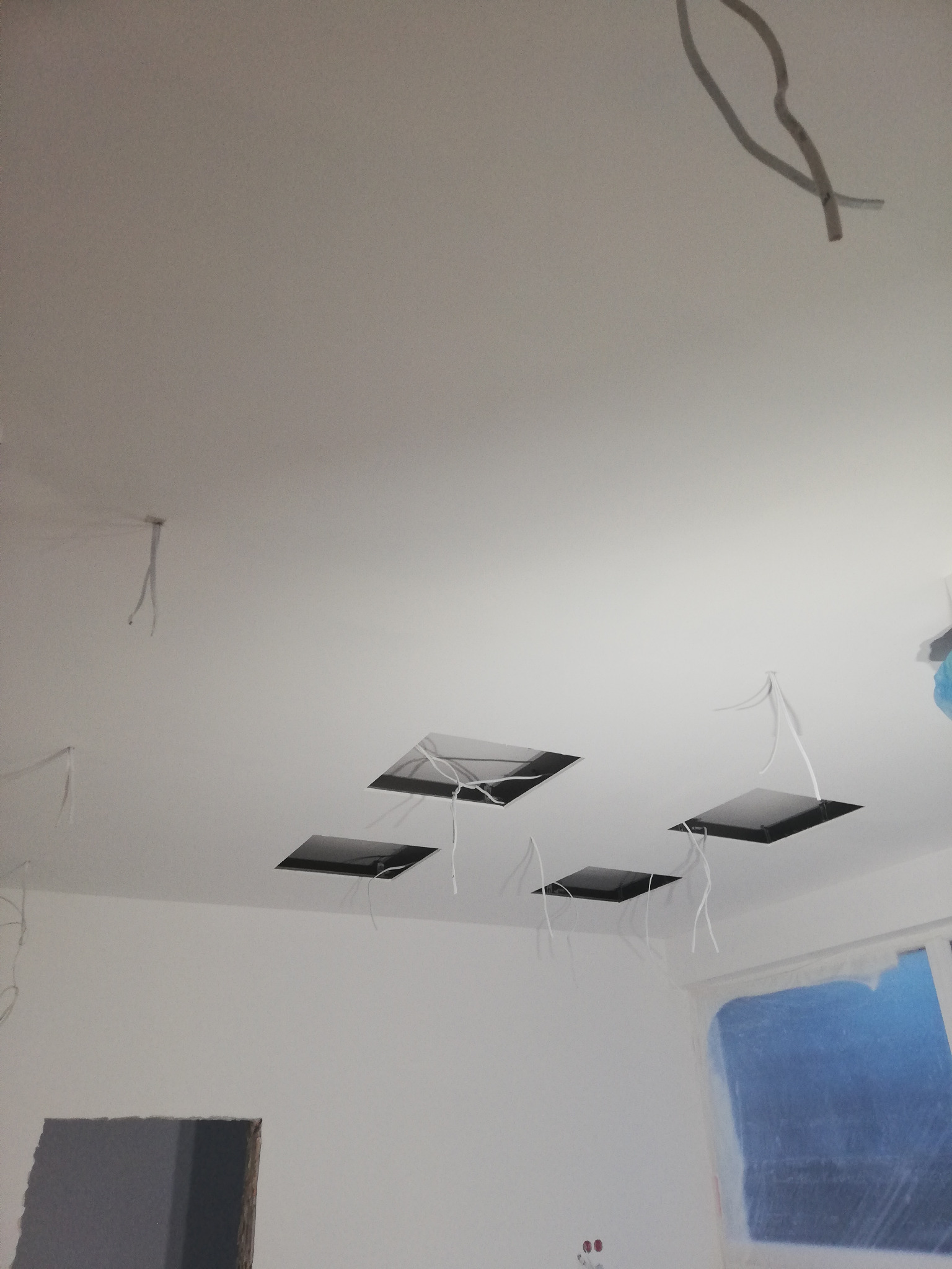 Biały sufit z czterema kwadratowymi otworami pod instalację oświetleniową, z których zwisają przewody, widoczne okno zabezpieczone folią malarską, ściany w trakcie malowania.