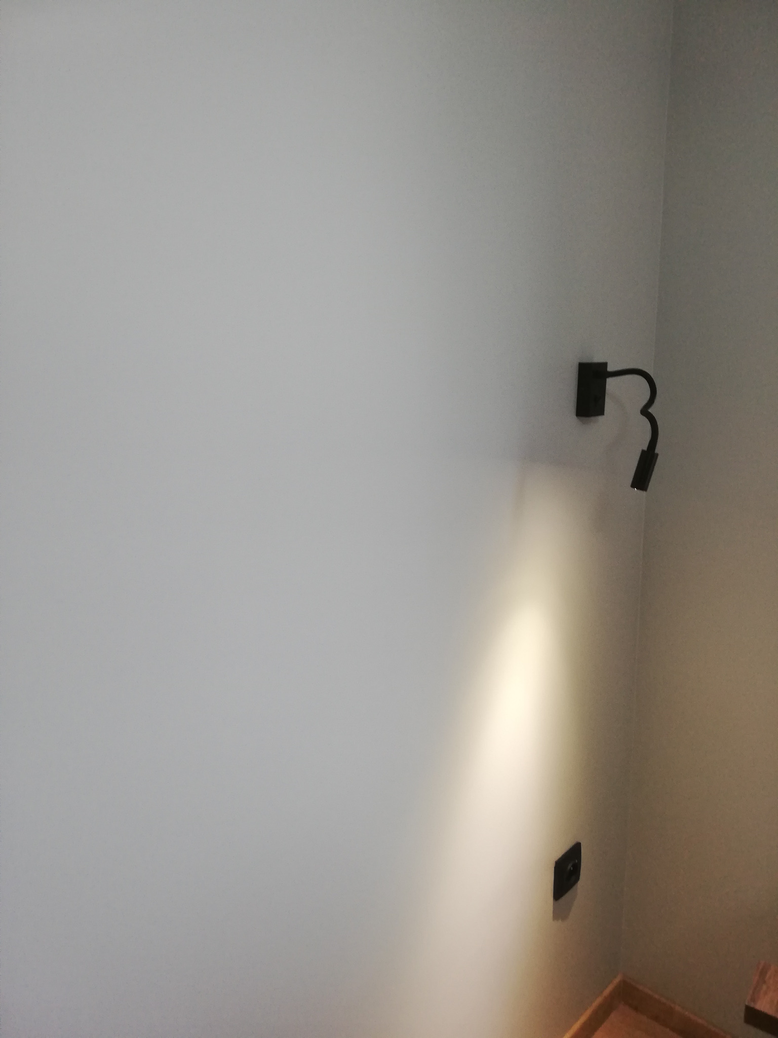 Szary narożnik pokoju z zamontowanym czarnym kinkietem punktowym i gniazdkiem elektrycznym, oświetlonym snopem światła.