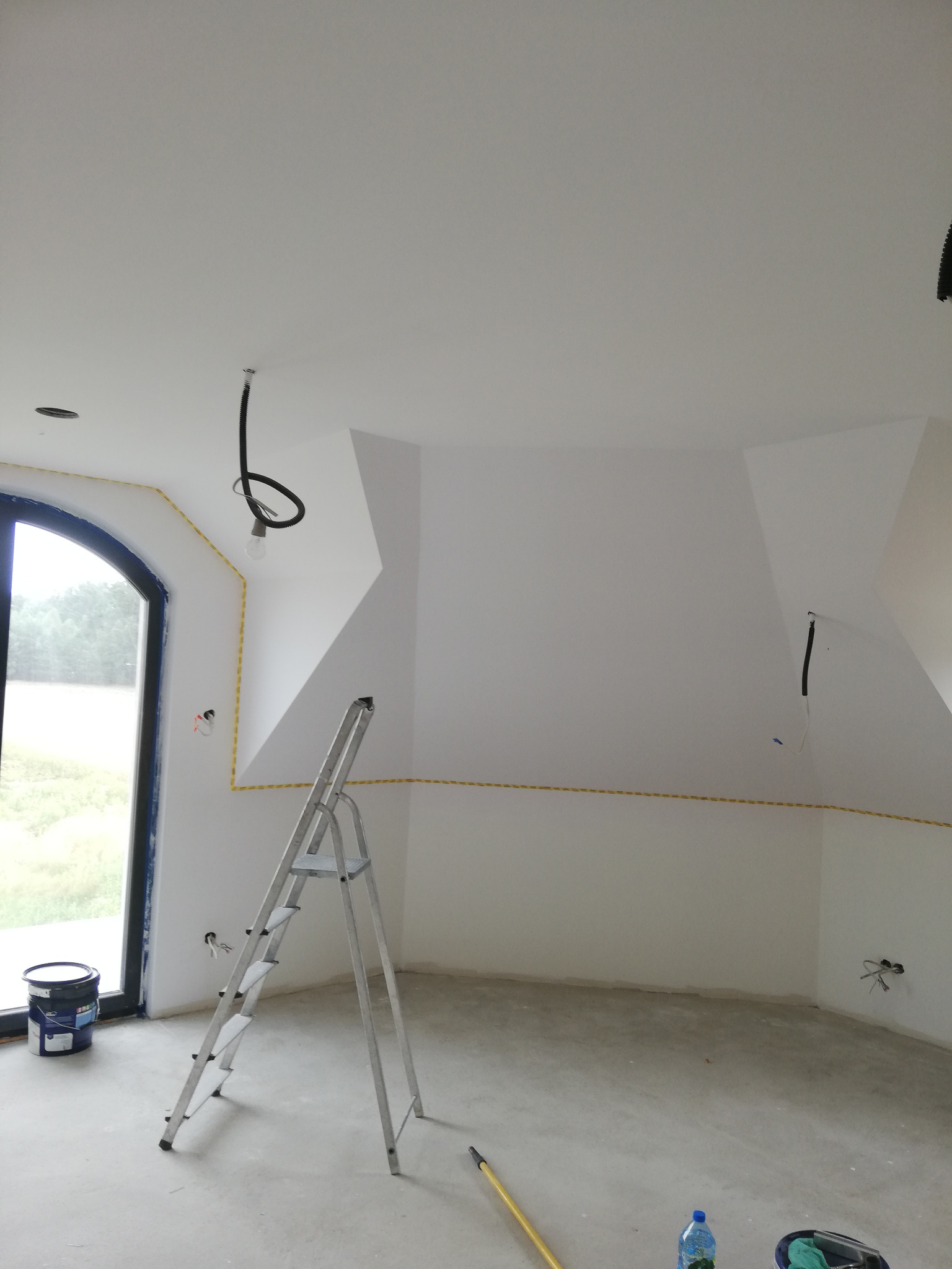 Wnętrze w trakcie malowania: białe ściany, surowa podłoga, aluminiowa drabina, puszka farby przy oknie, kable elektryczne zabezpieczone taśmą malarską