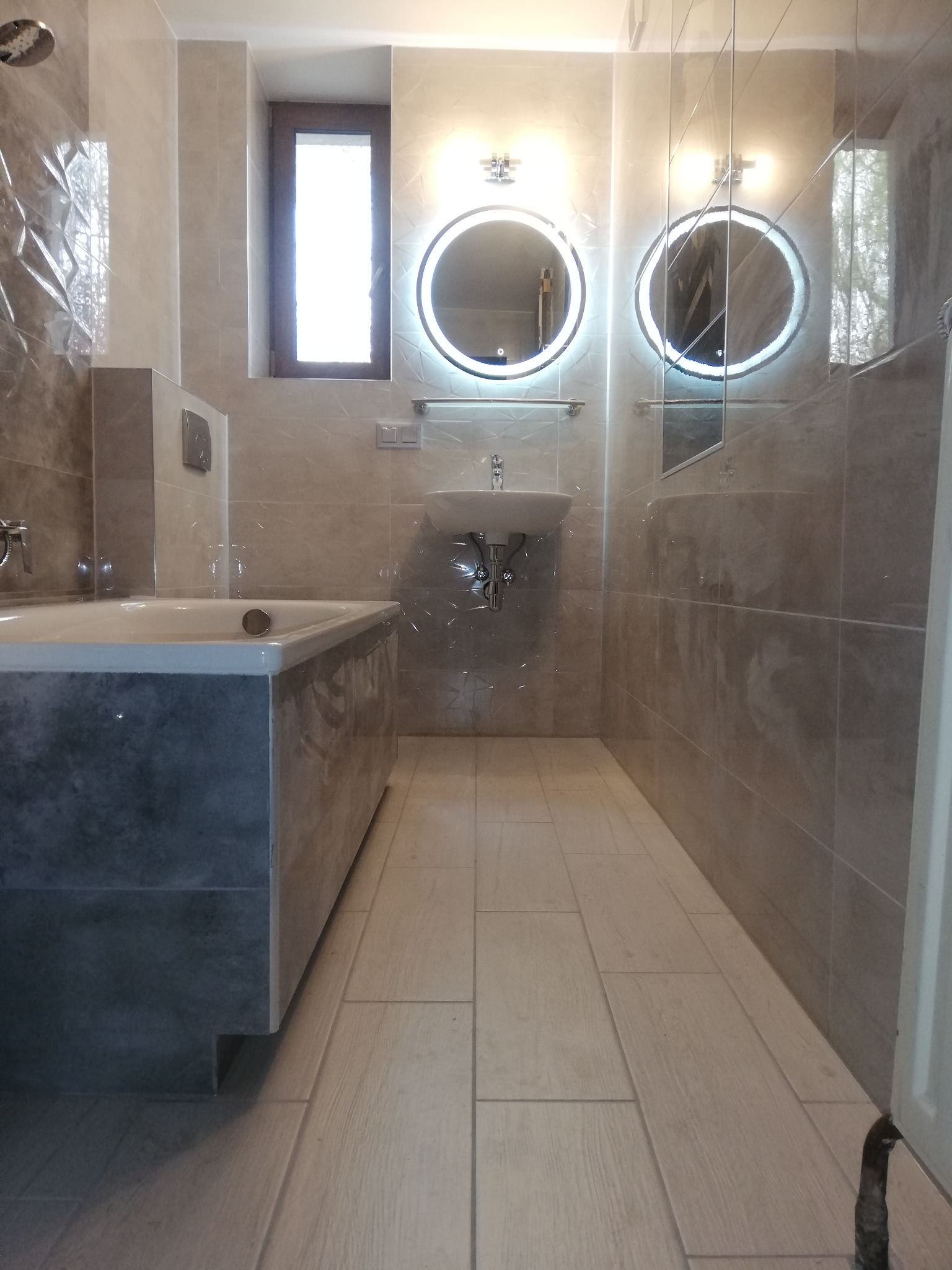 Nowoczesna łazienka z beżowymi płytkami, podświetlanymi okrągłymi lustrami, wanną obudowaną płytkami w odcieniach szarości i jasną podłogą z płytek imitujących drewno.