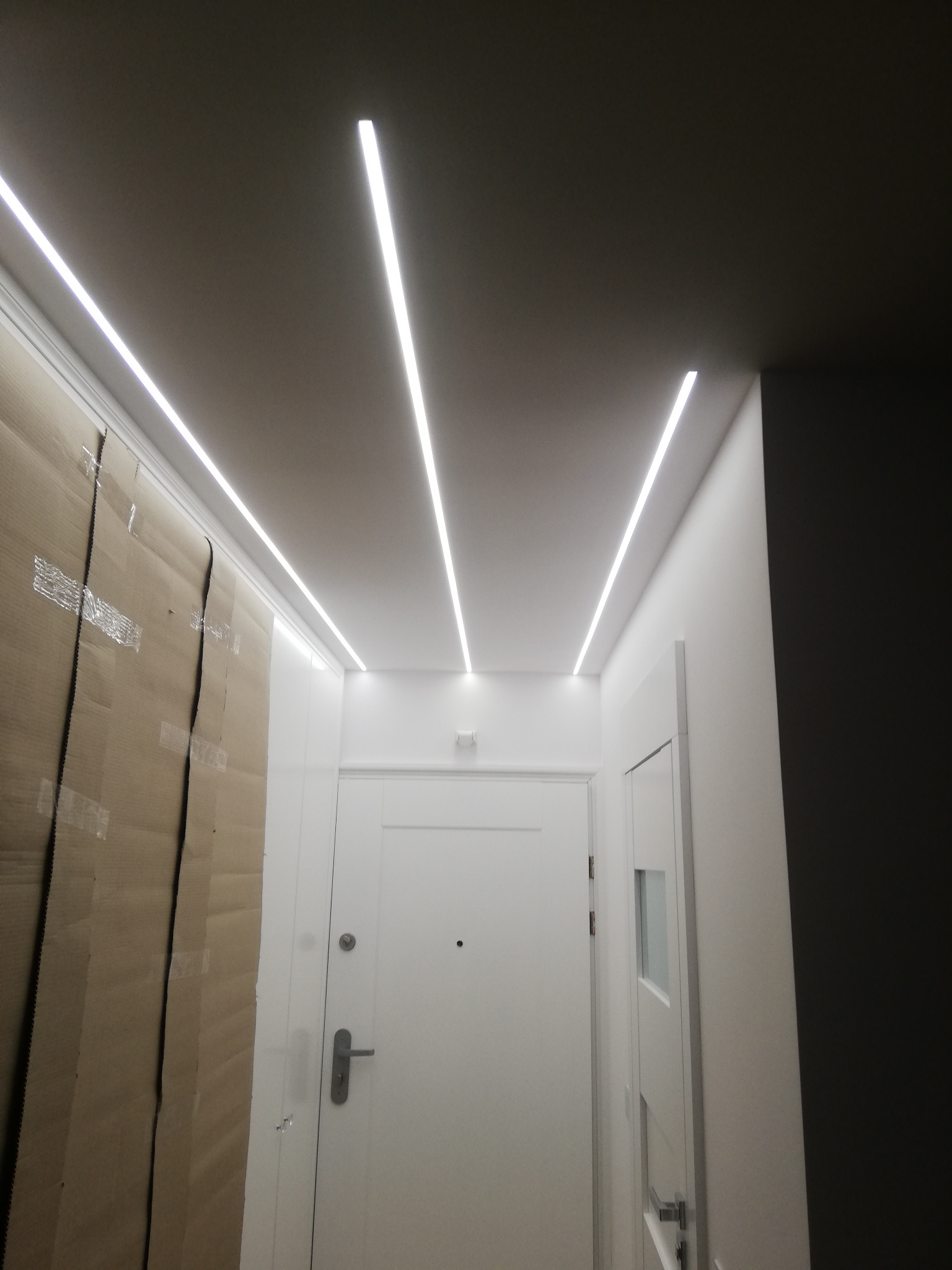Nowoczesne oświetlenie sufitowe LED w przedpokoju we Wrocławiu, z widoczną białą ścianą zabezpieczoną kartonem podczas remontu i białymi drzwiami.