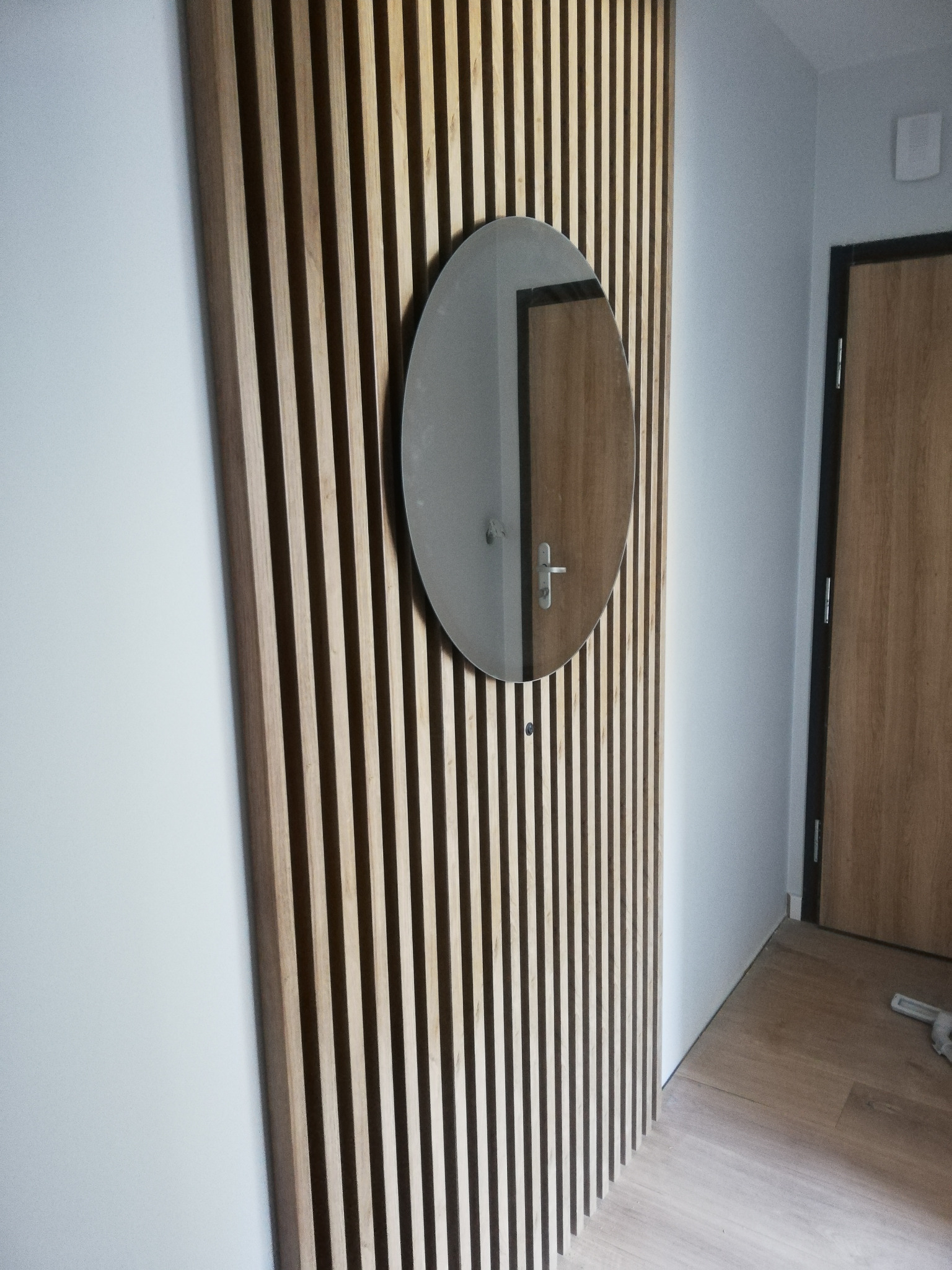 Drewniana ścianka działowa z pionowych listew z okrągłym lustrem, odbijającym drzwi, na tle jasnej ściany i podłogi.