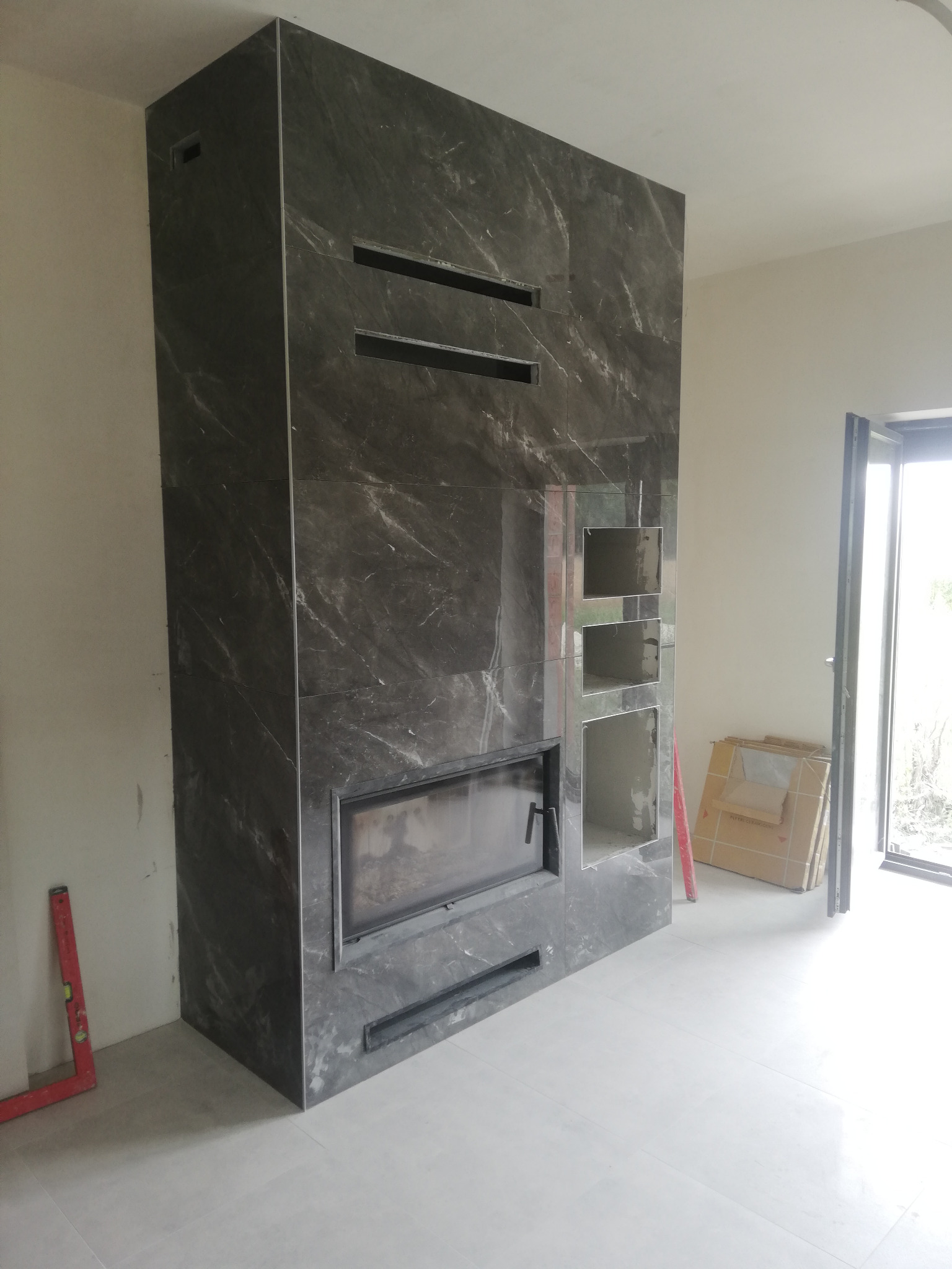 Obudowa kominka z szarego marmuru z czarnym wkładem kominkowym i wnękami, w trakcie montażu, poziomica oparta o ścianę, kartony z płytkami w tle.