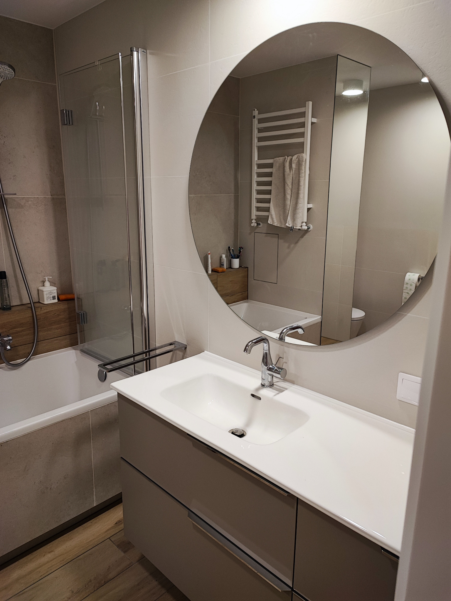 Nowoczesna łazienka z okrągłym lustrem, białą umywalką na szafce w kolorze taupe, kabiną prysznicową ze szklanymi drzwiami i grzejnikiem drabinkowym z ręcznikiem.