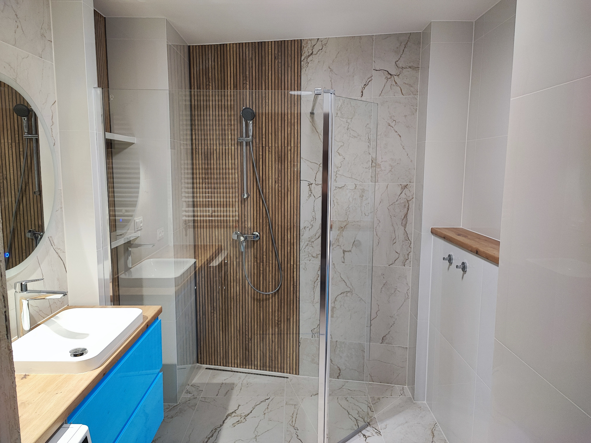 Nowoczesna łazienka z kabiną prysznicową typu walk-in, umywalką na szafce w kolorze turkusowym i płytkami imitującymi marmur na podłodze i ścianach, akcent z drewnianych paneli za prysznicem.