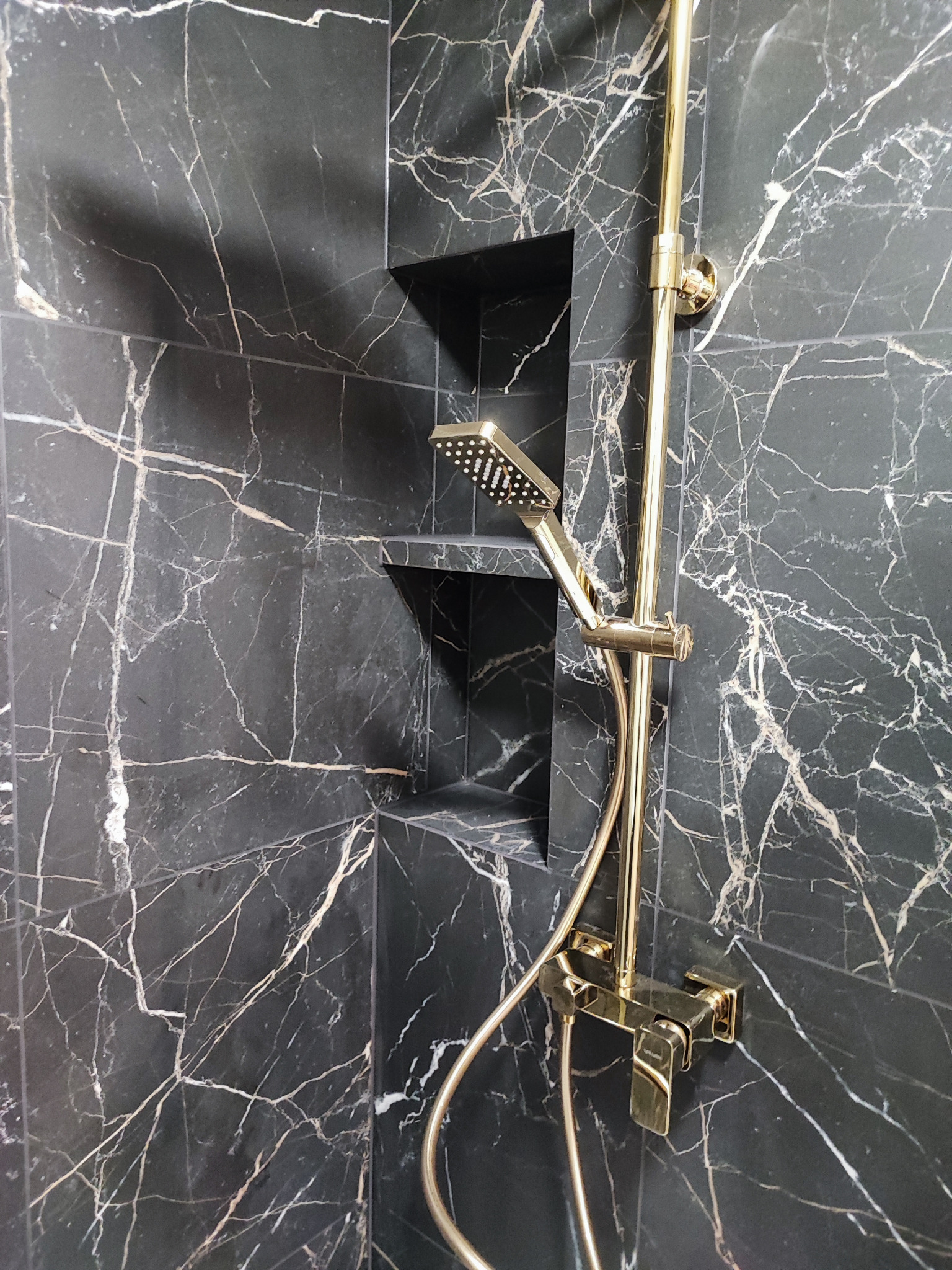 Bateria prysznicowa w kolorze złotym na tle ściany z czarnego marmuru z białymi żyłami oraz wnękami.