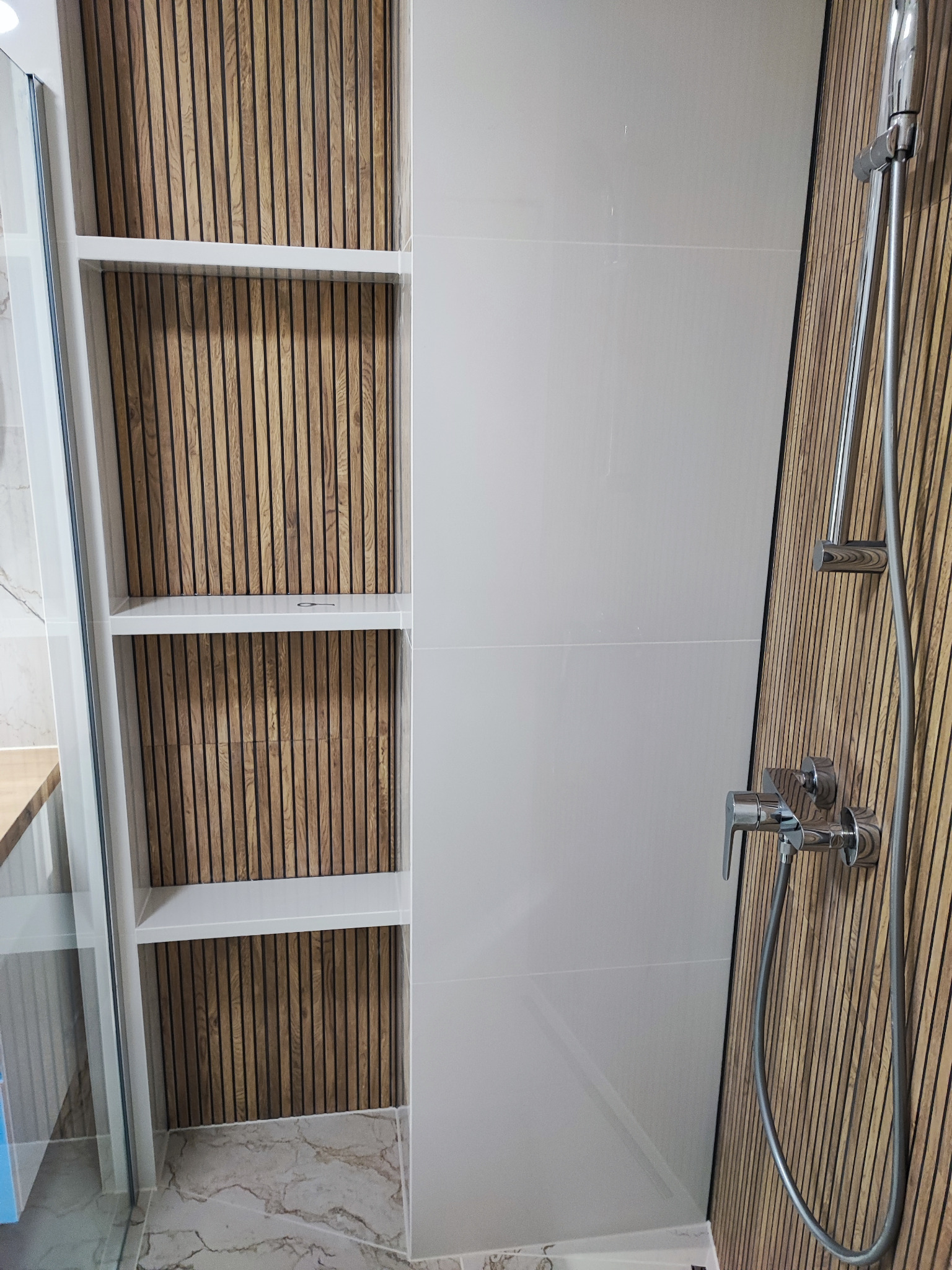 Nowoczesna łazienka z drewnianymi lamelami na ścianie, białe płytki, szklana kabina prysznicowa i chromowana armatura, widoczne półki w zabudowie.