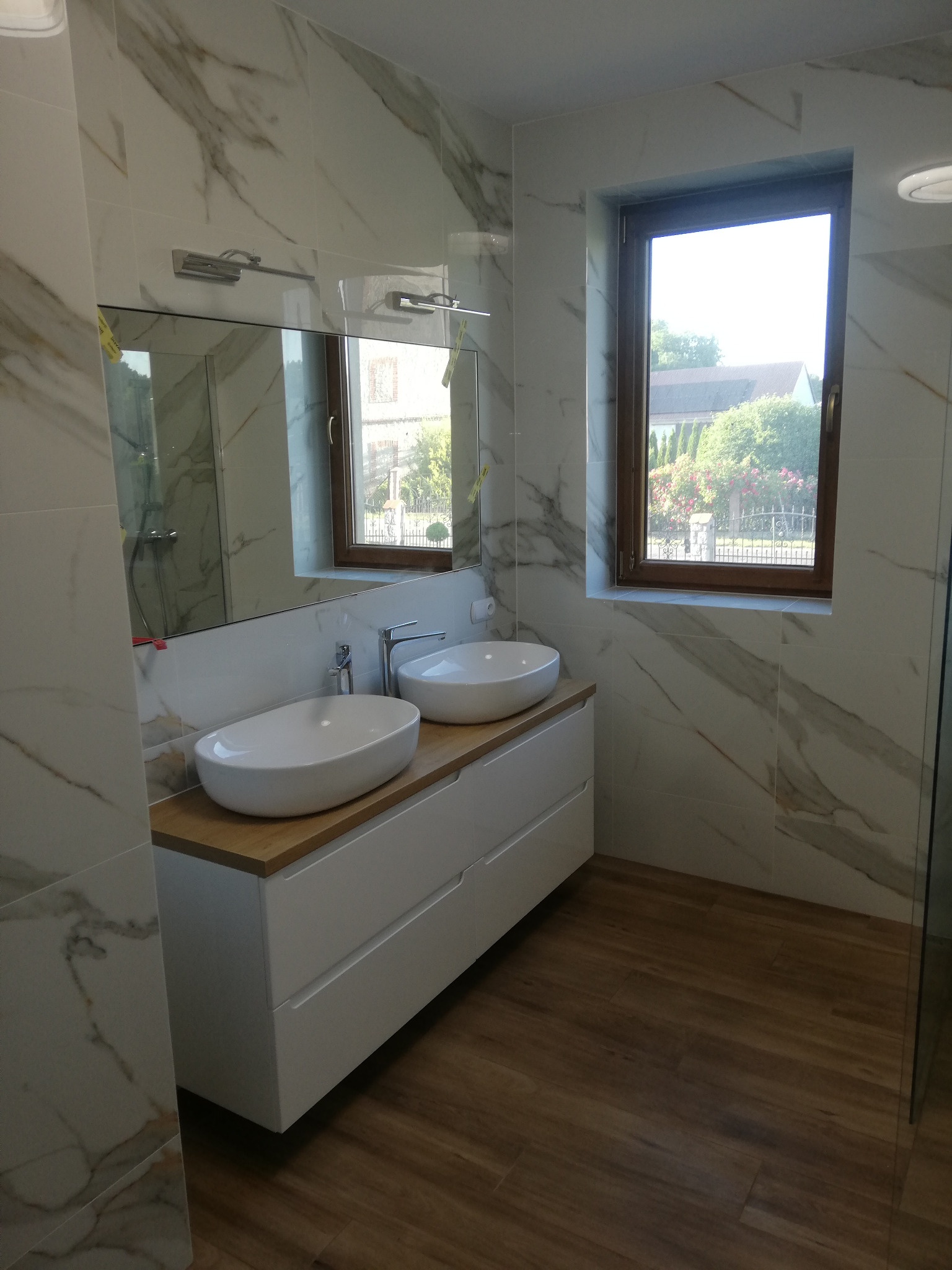 Nowoczesna łazienka z dwoma umywalkami nablatowymi, białą szafką pod umywalką z drewnianym blatem, marmurowe płytki na ścianach i drewnopodobna podłoga, okno z widokiem na ogród.