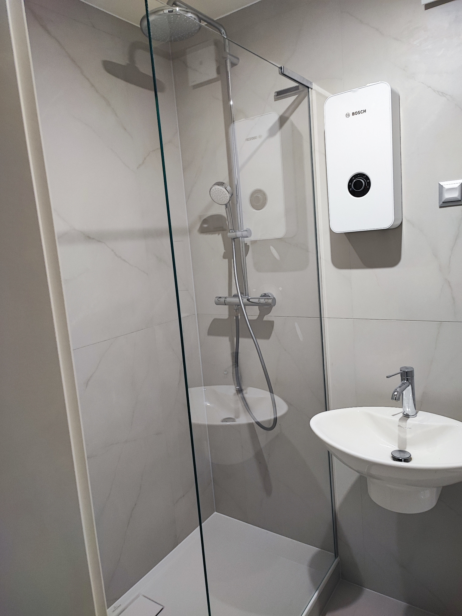 Nowoczesna kabina prysznicowa ze szklaną ścianą, deszczownicą i panelem prysznicowym, obok umywalka z baterią, białe marmurowe płytki na ścianach i podgrzewacz wody Bosch.