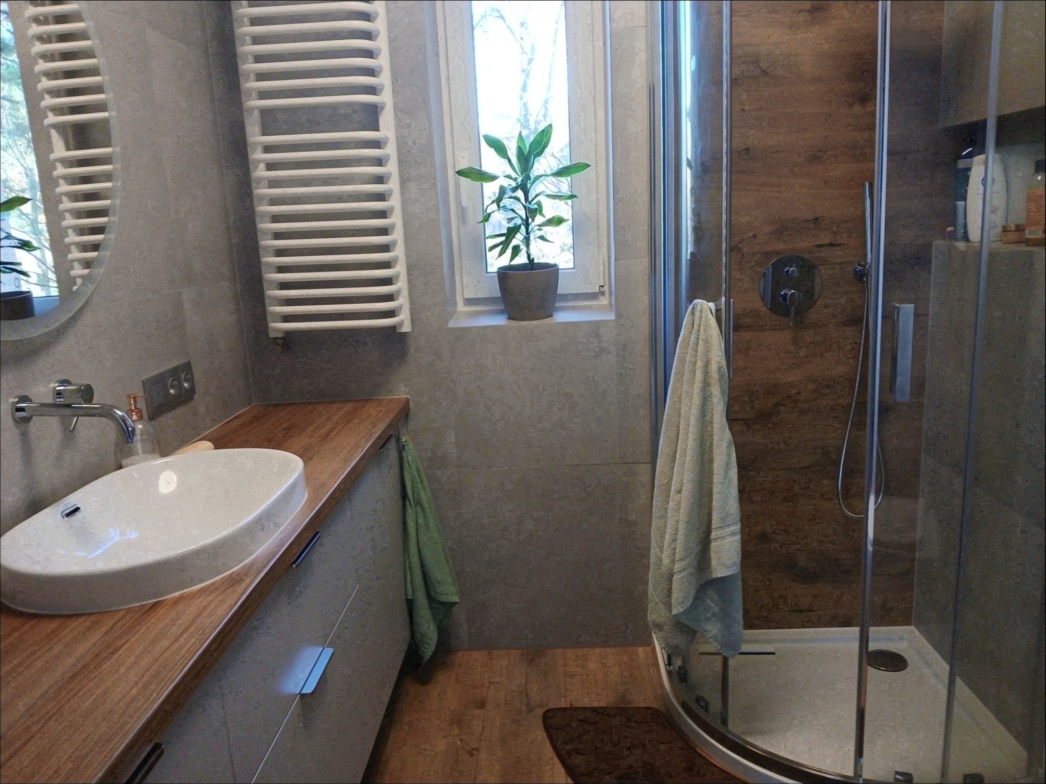 Nowoczesna łazienka z drewnianym blatem pod umywalką, grzejnikiem drabinkowym, kabiną prysznicową z drewnopodobnymi panelami i rośliną w doniczce na parapecie okna.