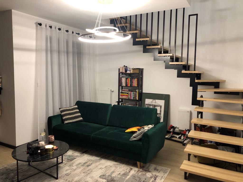 Nowoczesne schody drewniane z czarną metalową konstrukcją w jasnym wnętrzu, widok z dołu, obok zielona sofa i stolik kawowy.