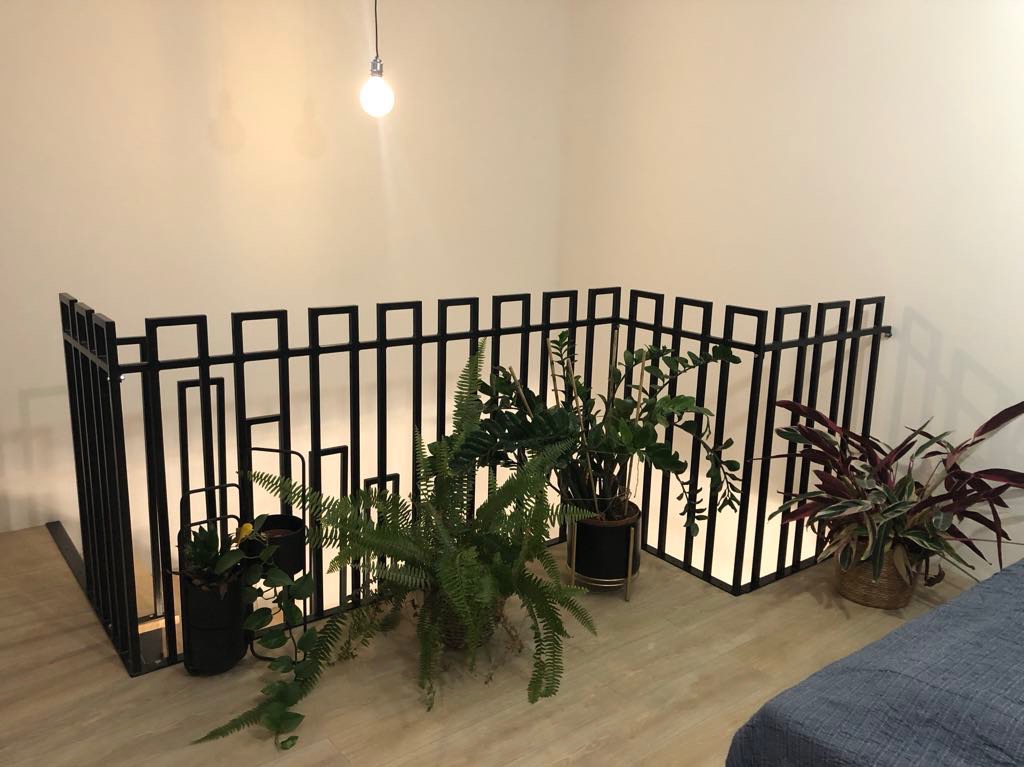 Czarna, metalowa barierka o nowoczesnym designie z kwadratowymi elementami, otoczona roślinami doniczkowymi w jasnym wnętrzu.