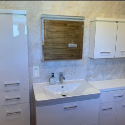 Firma Remontowo Budowlana SOLO Przemysław Kuszyński - Białe szafki łazienkowe z umywalką i lustrem na tle beżowych płytek. Nowoczesny design, chromowane uchwyty i bateria. Widoczny dozownik mydła i gniazdko.