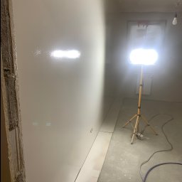 Firma Remontowo Budowlana SOLO Przemysław Kuszyński - Świeżo wygładzona ściana w trakcie prac wykończeniowych, oświetlona lampą budowlaną. Widoczne ślady po szpachlowaniu i przygotowaniu do malowania.