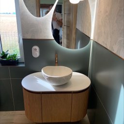 Firma Remontowo Budowlana SOLO Przemysław Kuszyński - Nowoczesna łazienka z umywalką w kształcie misy, podświetlanym lustrem o nieregularnym kształcie i drewnianą szafką. Widoczne zielone akcenty i tapeta w roślinny wzór.