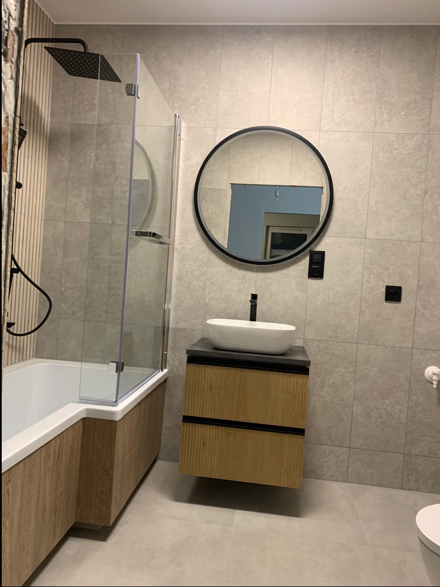 Nowoczesna łazienka z kabiną prysznicową typu walk-in, okrągłym lustrem i szafką pod umywalką w odcieniach drewna. Szare płytki na ścianach i podłodze.